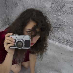 image of kneeling girl in a red dress looking at viewer and holding a camera. dark curls of hair obscure part of her face