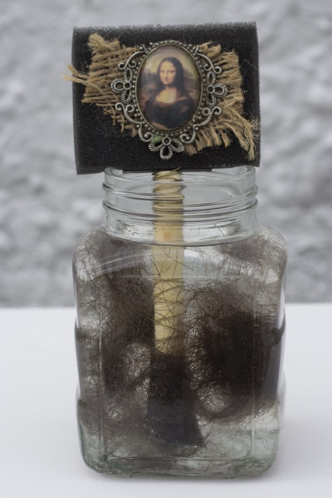 image of jar holding hair, with sign made from a paintbrush holding a cameo image of Mona Lisa pinned to burlap