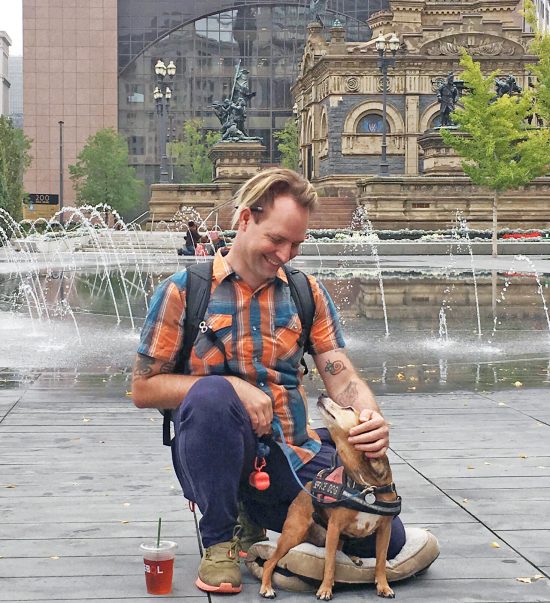 image of kneeling white-skinned man with small service dog in front of a fountain